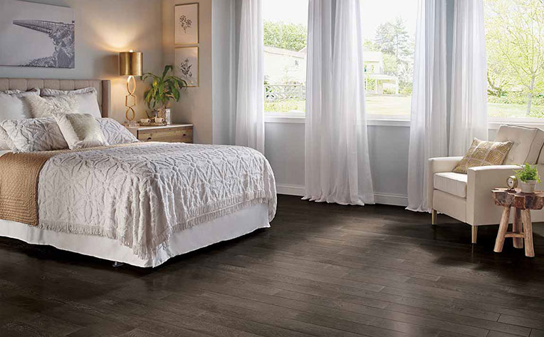 Bedroom with a bed covered white and gold bedspread on wood floors with white curtains.
